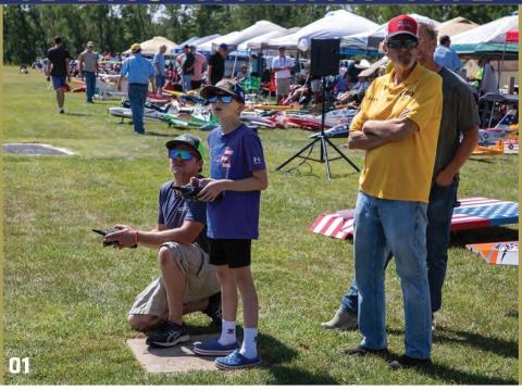 A young RC pilot flies under guidance from his father at an RC club air show, with another club member standing nearby.