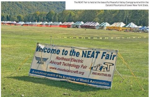 Sign welcoming visitors to the NEAT Fair in a grassy field with rows of tents and wooded mountains behind it.